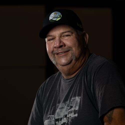 J.B. Hunt driver smiling for a dimly lit headshot picture wearing a J.B. Hunt graphic tee and hat.