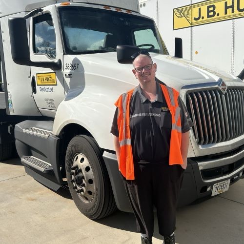 Woman J.B. Hunt driver stands in front of three J.B. Hunt Intermodal shipping containers with a smile on her face. She is wearing a hat with a J.B. Hunt logo on it and a bright colored safety vest