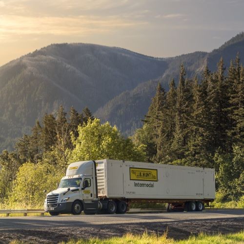 J.B. Hunt Driver wearing a cowboy hat, standing proudly with his arms crossed and a smile on his face. Behind him is a J.B. Hunt Intermodal container and a patriotic J.B. Hunt cab. The cab is patriotically wrapped with a flag design across the front hood and continues to the doors.