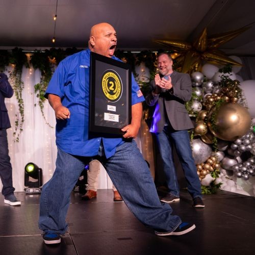 "J.B. Hunt driver, Adrian, is excitedly cheering on stage, while holding his two million collision-free miles plaque. Two men are cheering him on in the background standing next to balloon decorations.