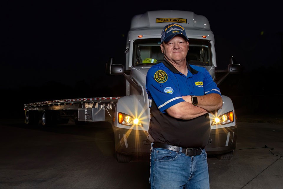 A J.B. Hunt truck driver standing confidently in front of a J.B. Hunt semi-truck at night, wearing a uniform with company logos, arms crossed, and illuminated by the truck’s headlights.