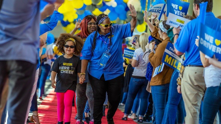A celebratory scene at J.B. Hunt’s headquarters featuring professional drivers and their families walking down a red carpet lined with cheering employees holding signs and balloons, recognizing drivers who have safely driven over a million miles.