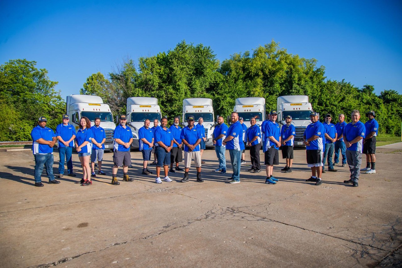 Group of drivers standing in front of 5 trucks in a parking lot.