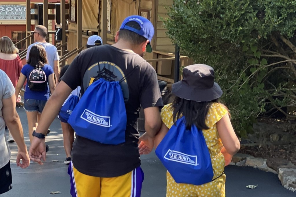 A group of people walking outdoors, with two individuals wearing blue J.B. Hunt drawstring bags, possibly attending a company event or gathering.