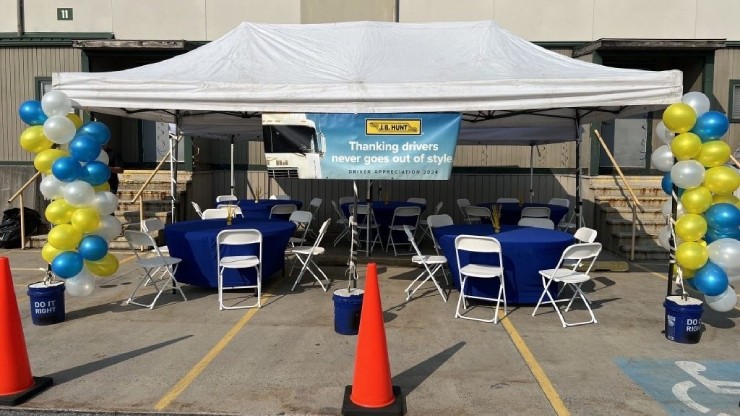A decorated outdoor tent setup with blue and yellow balloons, tables, and chairs for the J.B. Hunt Driver Appreciation 2024 event, featuring a banner that reads “Thanking drivers never goes out of style.”