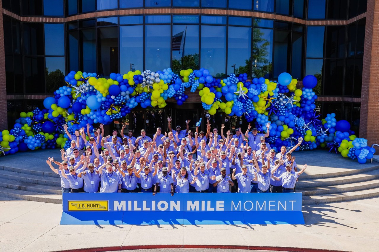 A group of Million Mile drivers standing in front of the J.B. Hunt corporate office with arms up behind a sign that reads "Million Mile Moment".