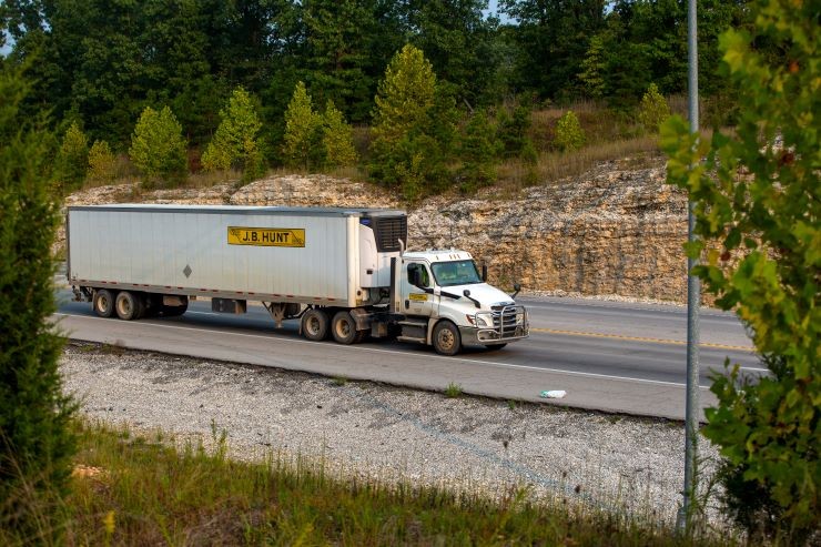 J.B. Hunt Semi-Truck Driving on a Highway A J.B. Hunt semi-truck hauling a refrigerated trailer on a highway, surrounded by trees and rocky terrain.