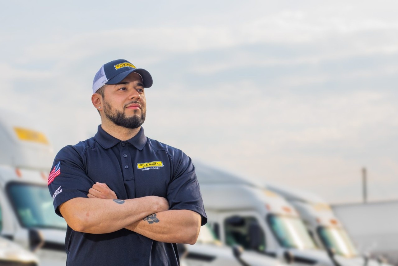 J.B. Hunt Intermodal driver standing with arms crossed in front of several J.B. Hunt branded trucks.