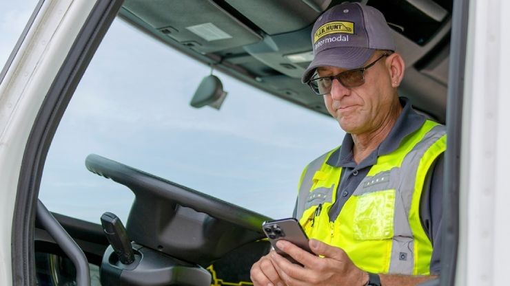 Truck driver in safety vest and cap sitting in cab using mobile phone to access job transfer portal for new driving opportunities.