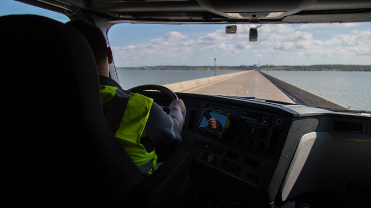 Truck Driver Crossing a Bridge with a Scenic Water View Truck driver wearing a high-visibility vest driving across a long bridge over water under a partly cloudy sky.