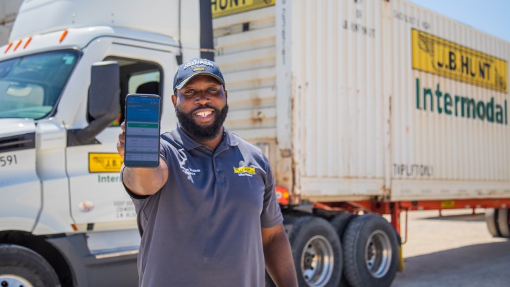 A smiling J.B. Hunt truck driver holds up a smartphone displaying a logistics app, standing in front of a semi-truck and intermodal container.