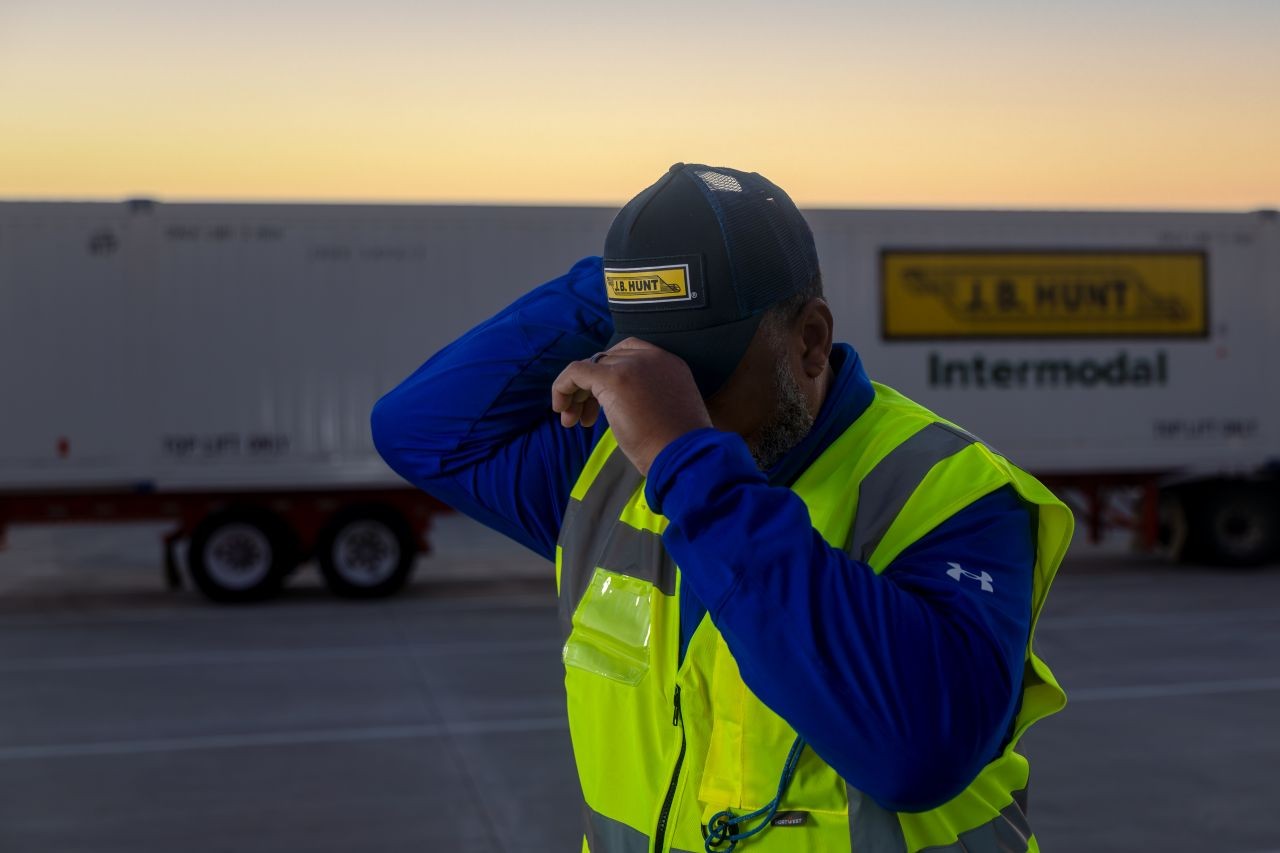 A driver wearing a high visibility vest pulls on a hat while standing in front of a J.B. hunt intermodal trailer