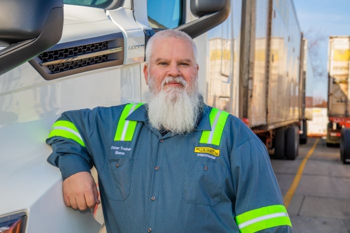 A professional truck driver trainer with a long white beard and a confident expression leans against a semi-truck, wearing a J.B. Hunt uniform with reflective safety stripes.