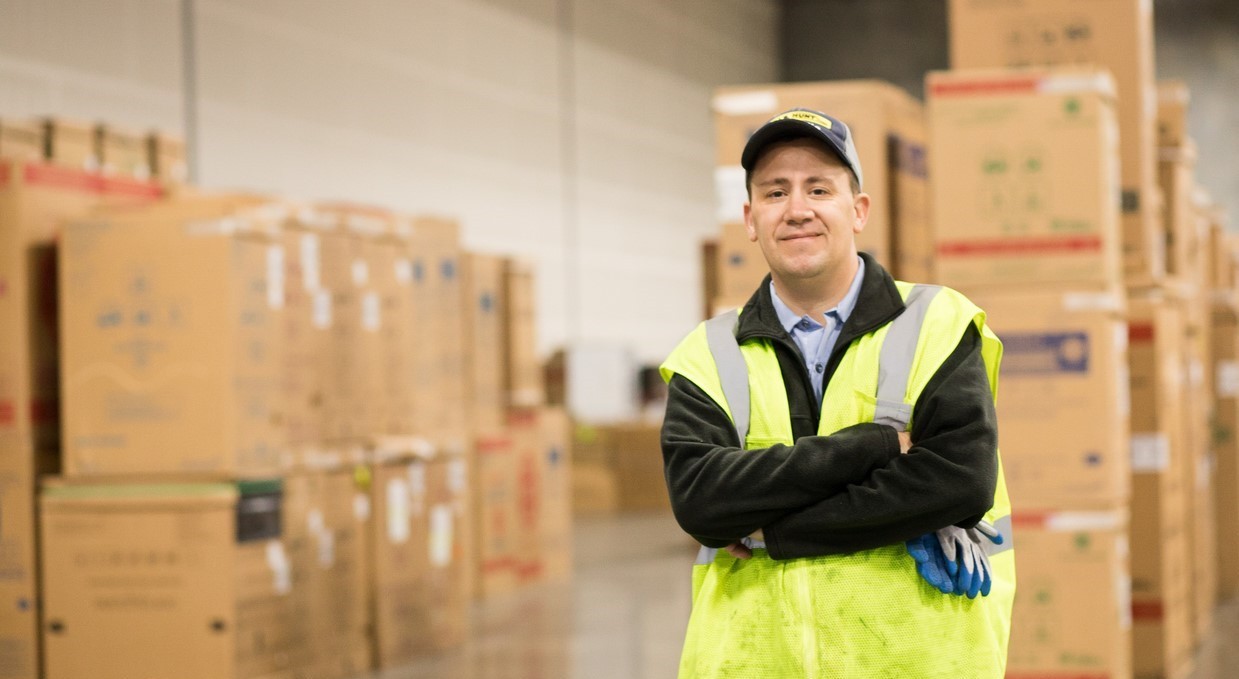 J.B. Hunt Final Mile Services driver John stands with arms crossed in front of warehouse boxes.