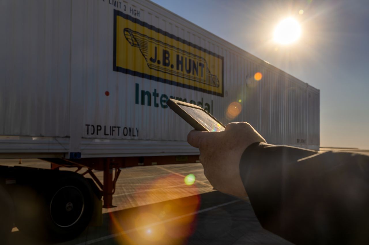 Person using a mobile device in bright sunlight near a J.B. Hunt intermodal container on a trailer, with lens flare highlighting modern freight technology.
