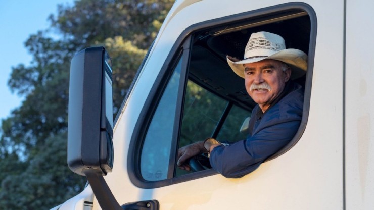 A smiling older truck driver wearing a cowboy hat leans out of the cab of his semi-truck, looking at the camera with a friendly expression.
