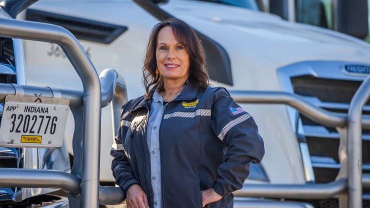 Confident Female Truck Driver Standing in Front of Semi-Trucks A female truck driver wearing a company jacket stands confidently in front of two large semi-trucks, with a truck license plate visible in the foreground.