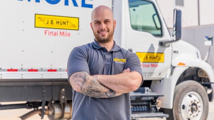 Marcus, a delivery driver with a beard and tattoos, wearing a J.B. Hunt Final Mile uniform, stands confidently with arms crossed in front of a company delivery truck.
