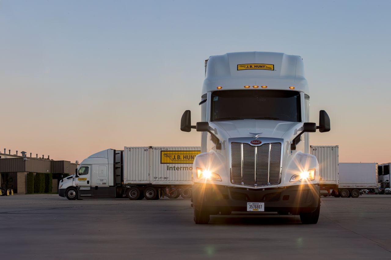 J.B. Hunt semi-truck with headlights on, parked in a lot with intermodal containers at sunset.