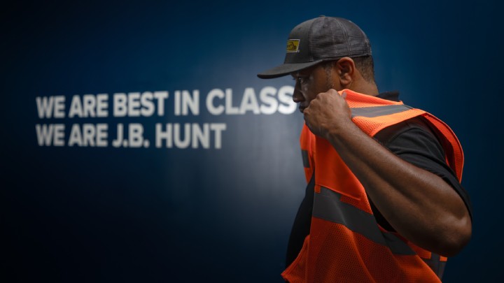 A person wearing an orange safety vest and a dark cap adjusts the vest while standing in front of a blue wall with white text.