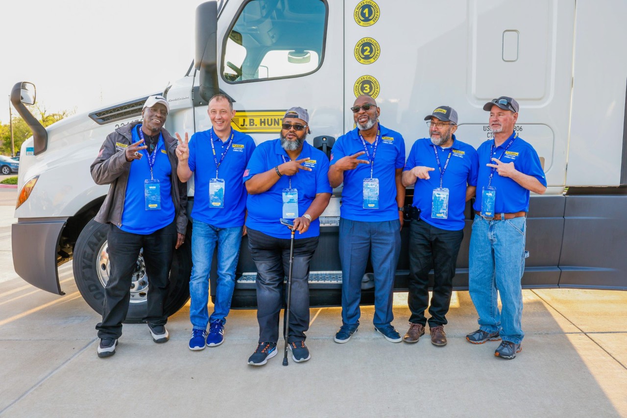 6 J.B. Hunt Drivers smiling and holding up two fingers while standing in front of a J.B. Hunt truck