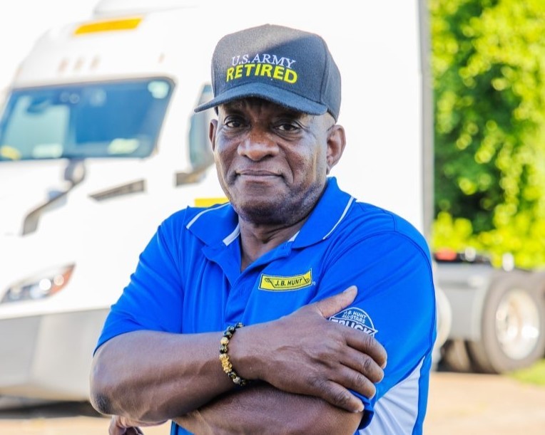 A retired U.S. Army veteran wearing a “U.S. Army Retired” cap and a blue J.B. Hunt uniform stands confidently with arms crossed in front of a semi-truck.
