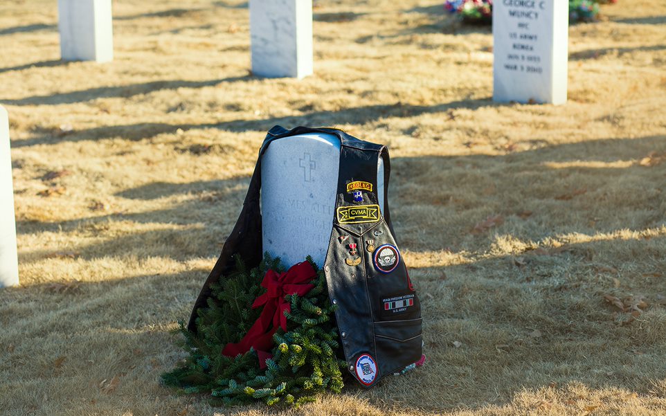 Green wreath laid in front of grave stone dressed in leather military-patched vest.