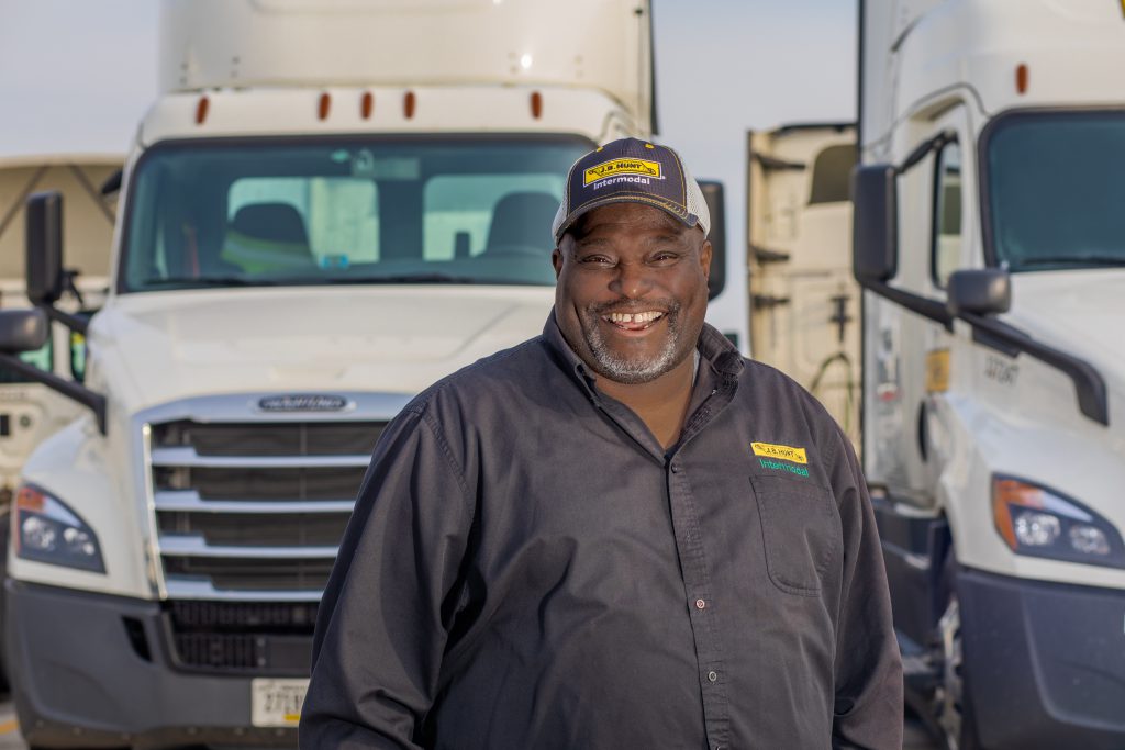 Driver Kenneth stands in front of two J.B. Hunt Intermodal trucks smiling.