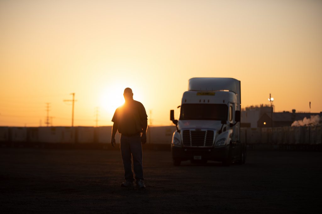 Driver silhouette standing in front of a truck with the sun shining behind him.