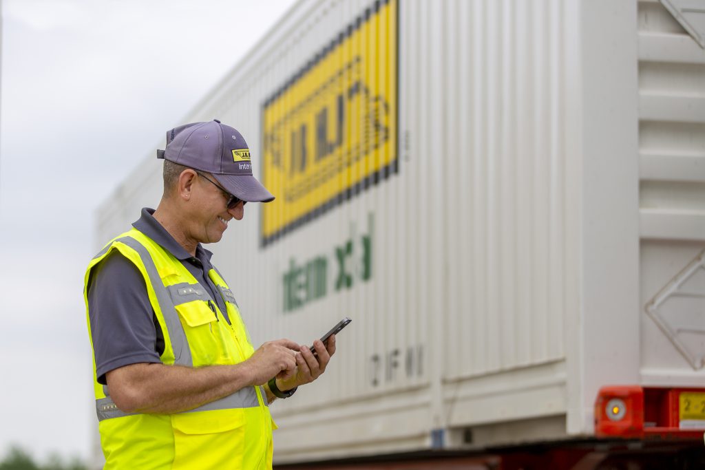 J.B. Hunt driver stands in front of an intermodal container, looking at phone and smiling.