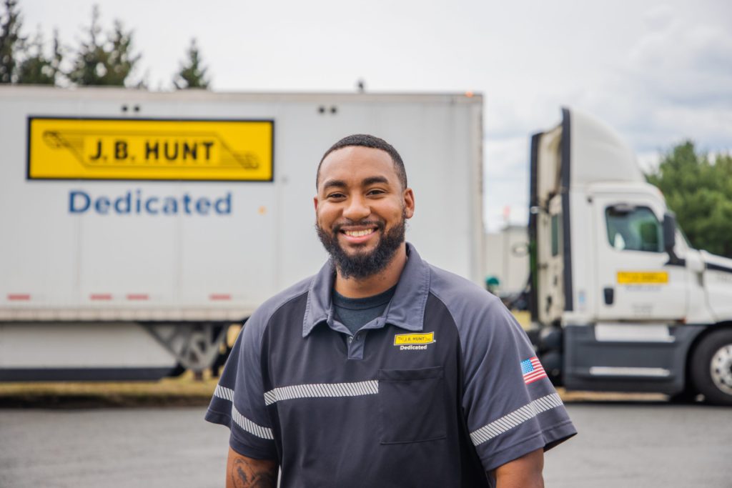 J.B. Hunt driver stands in front of a J.B. Hunt day cab and trailer. Smiles.
