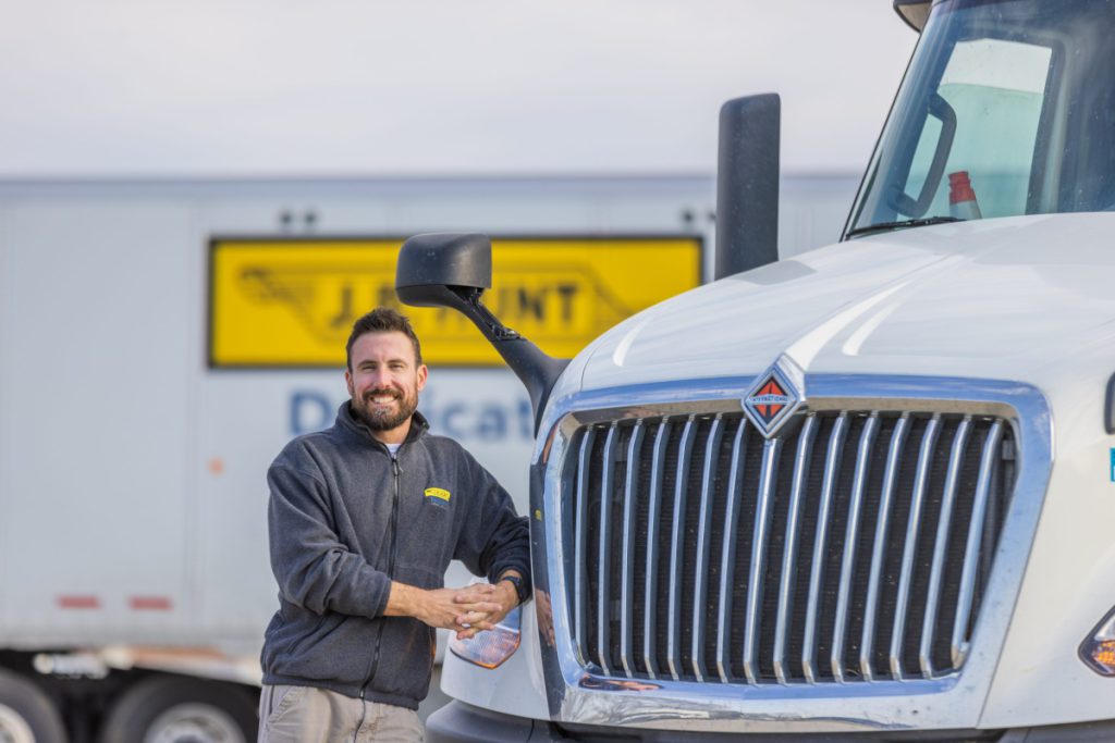 J.B. Hunt driver leans against a J.B. Hunt day cab. J.B. Hunt Dedicated Contract Services® trailer in the background.