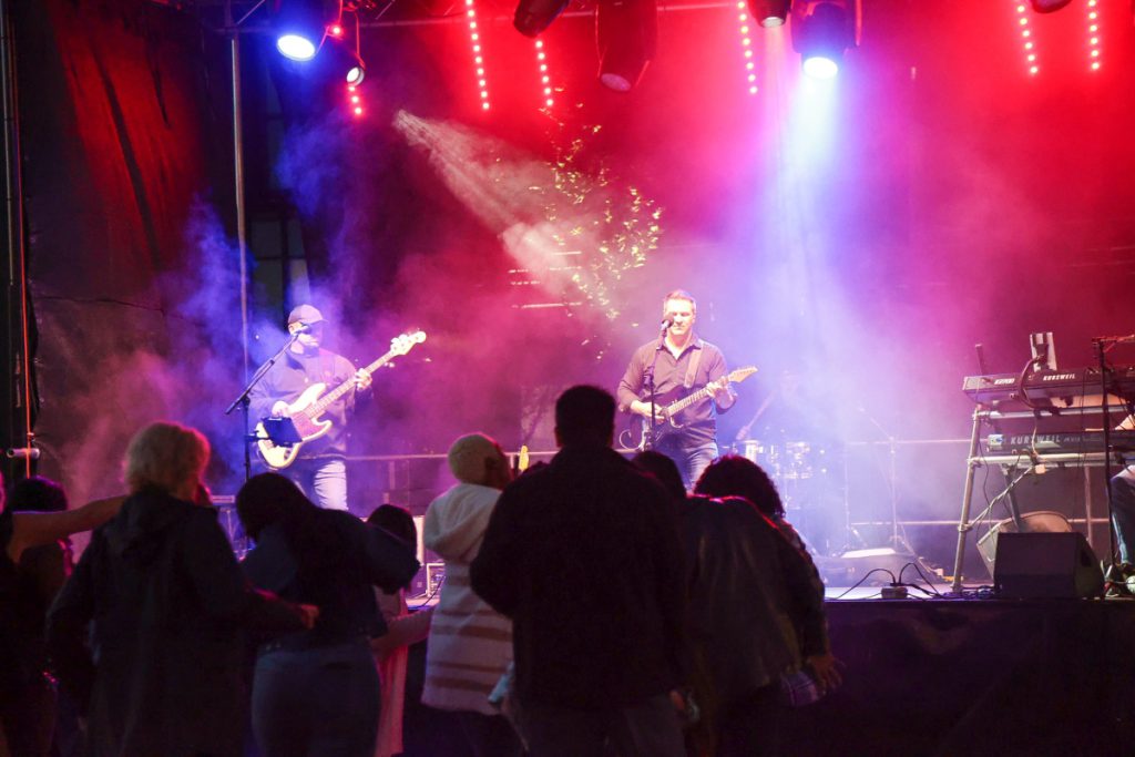 A band plays at night under stage lighting, people dancing to the music.