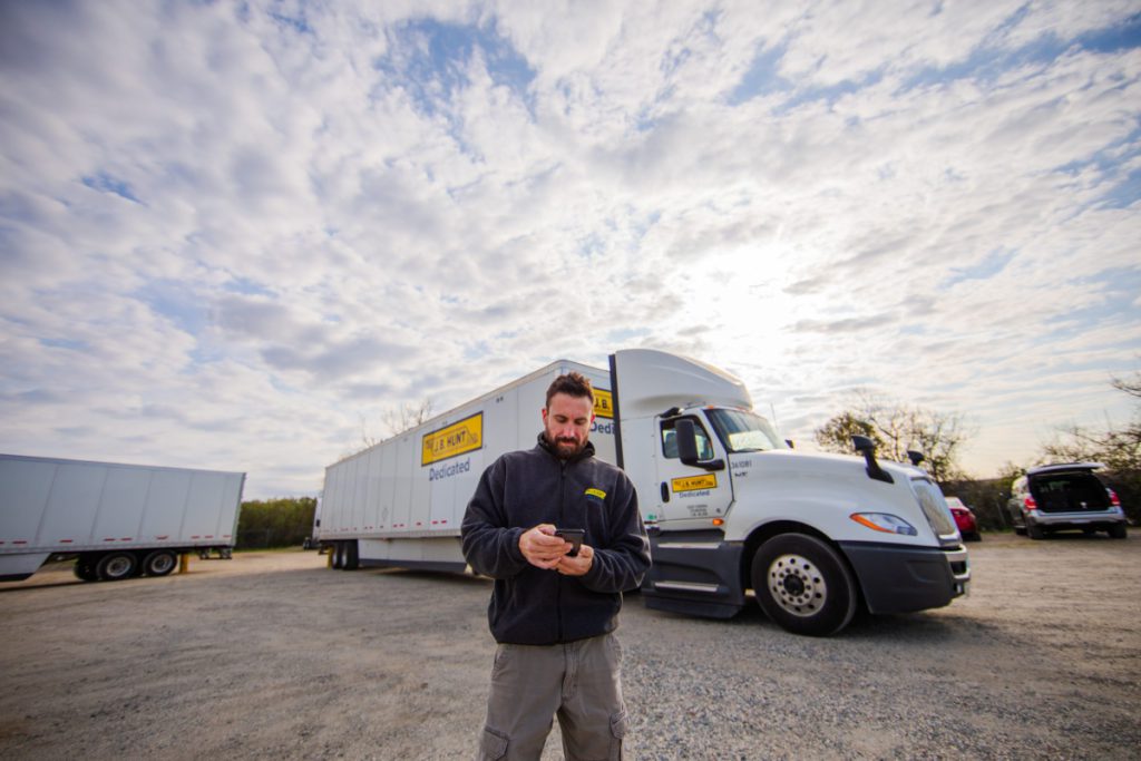 J.B. Hunt driver stands in front of a parked tractor trailer and checks the DRIVE App.