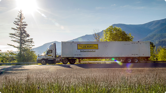 A white J.B. Hunt Intermodal semi-truck with a container trailer drives along a scenic mountain road under a bright, sunny sky with lens flare. Lush green trees and distant mountains frame the background, while tall grass lines the foreground.