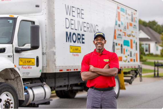 A smiling J.B. Hunt Final Mile delivery driver in a red polo shirt and cap stands proudly in front of a white straight truck. The truck, marked 'WE'RE DELIVERING MORE,' signifies J.B. Hunt's commitment to delivering and installing appliances, furniture, and other products to customers' homes and businesses. This image represents the fulfilling career opportunities available for non-CDL straight truck drivers and Delivery Installation Specialists at J.B. Hunt, with no experience required.