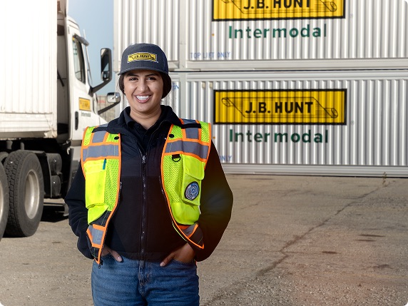 Smiling woman in a J.B. Hunt cap and safety vest, representing new and student drivers, standing confidently with a semi-truck and J.B. Hunt Intermodal containers in the background. This image highlights J.B. Hunt driving jobs for students and new professionals in the intermodal sector.